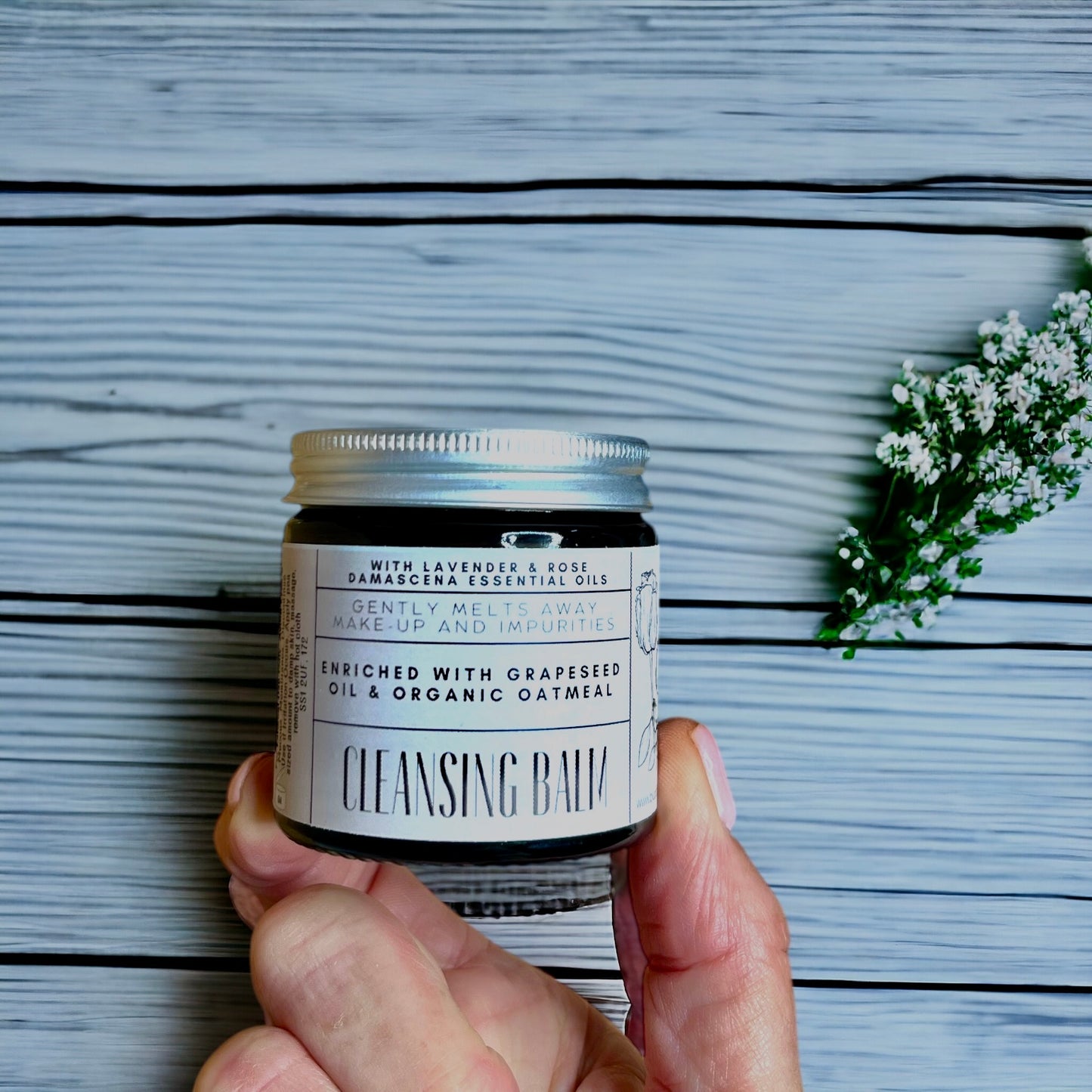 featuring a hand holding an amber glass jar of cleansing balm made with oats and lavender and rose Damascena essential oils made by buddha and bubbles botanicals