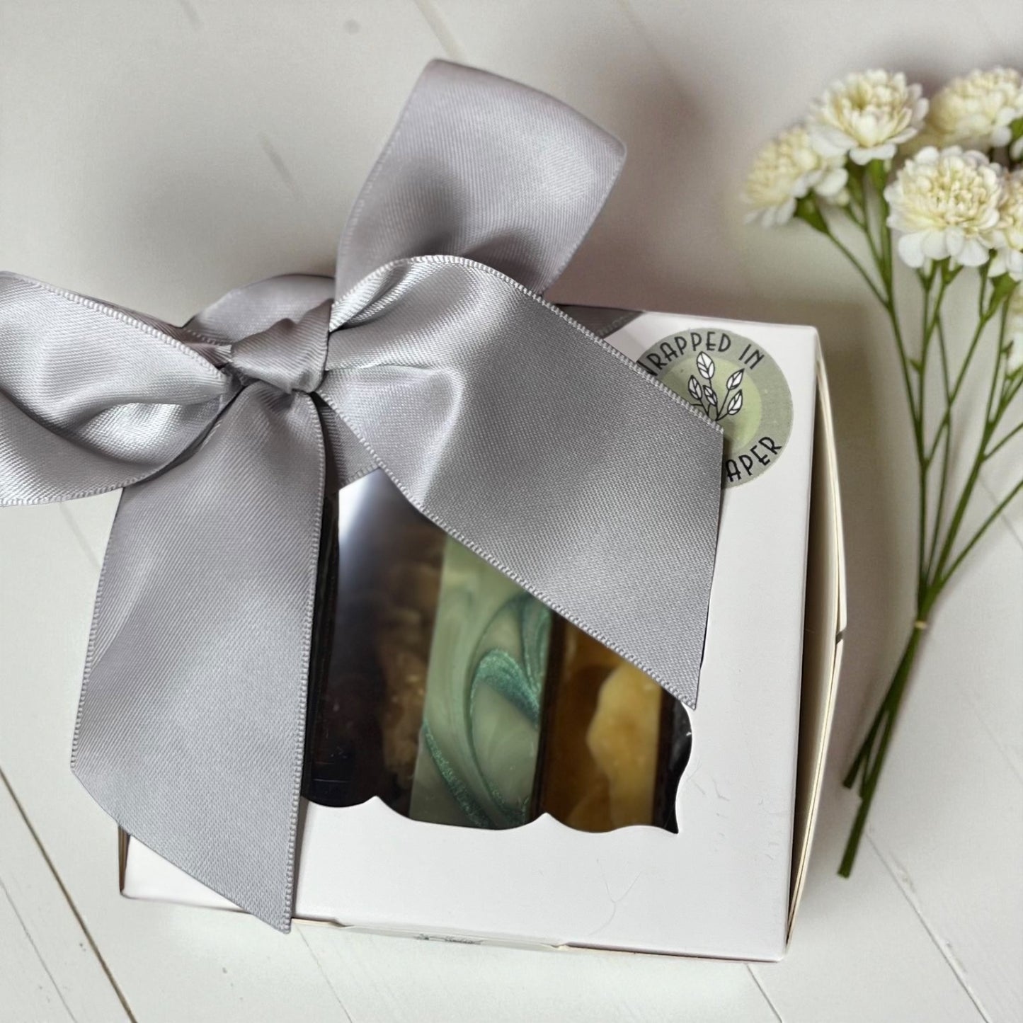 six small soaps in a box with a beige ribbon, next to white flowers on a light surface.
