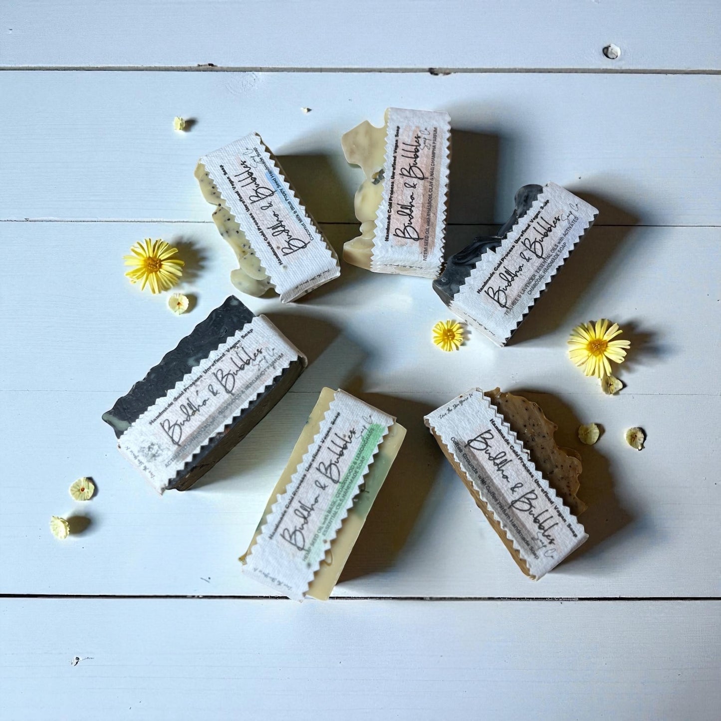 Collection of six small soap bars with visible branding on a light wooden surface with small yellow flowers.