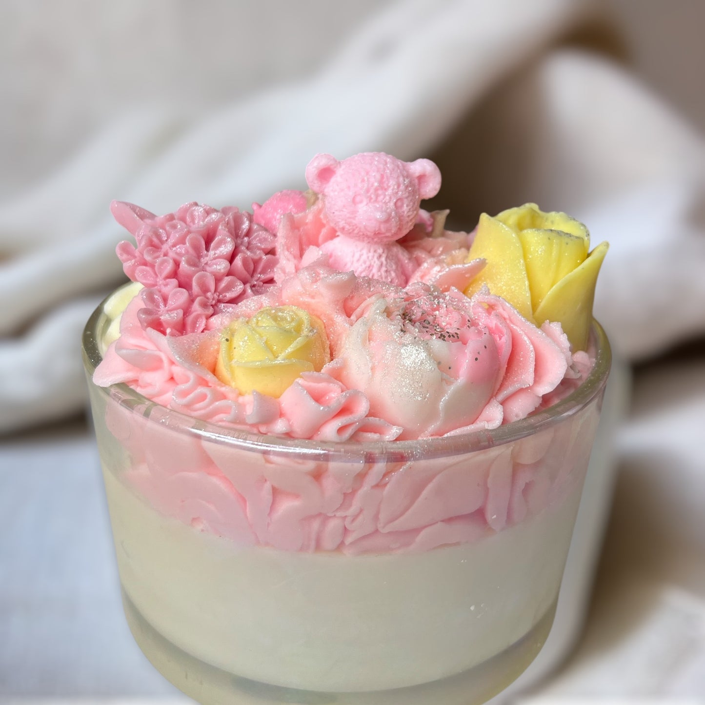 Decorative glass candle with pink whipped wax, flowers, and a small bear on a white table