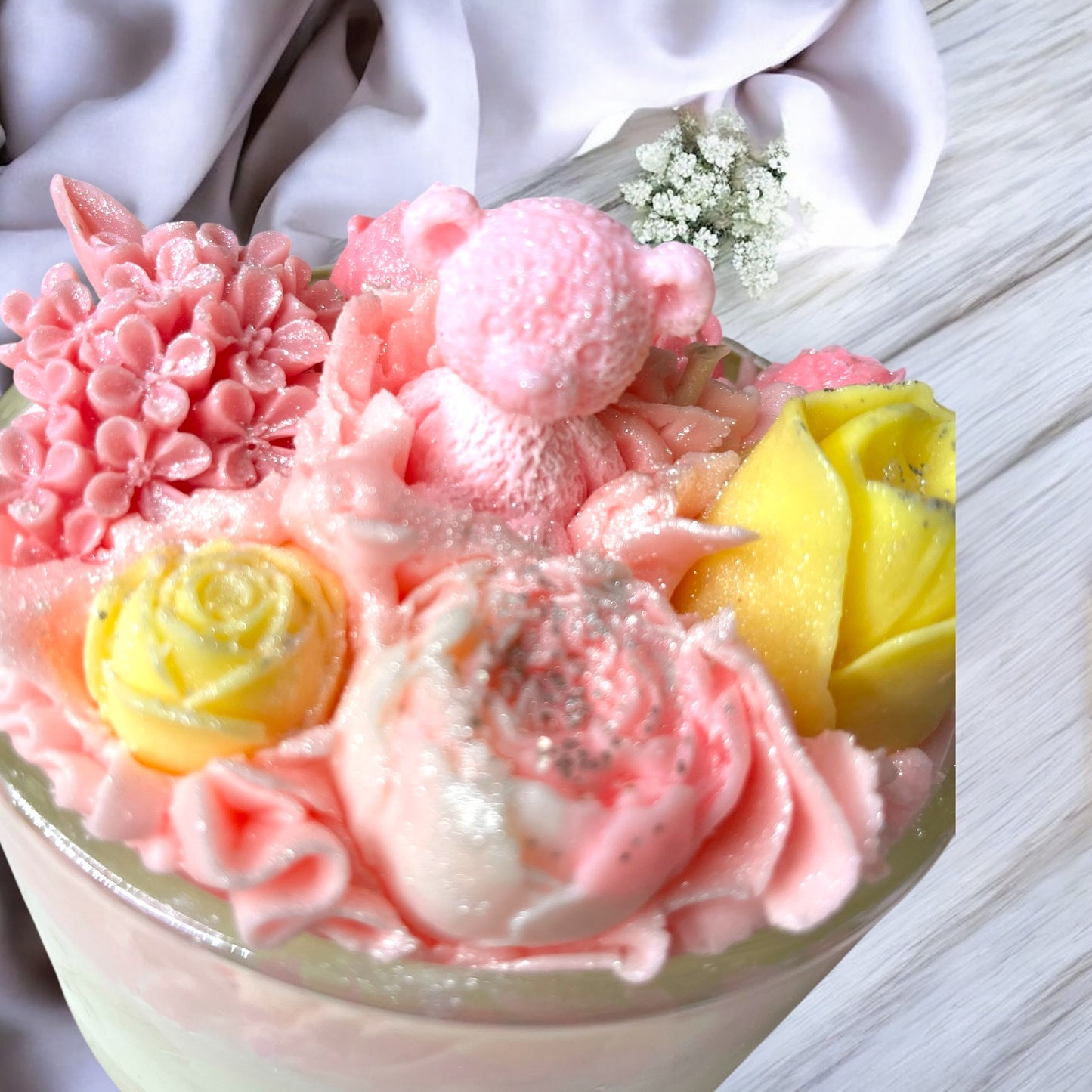 large botanical candle with pink and yellow flowers and a teddy bear in glass container on a light wooden surface.