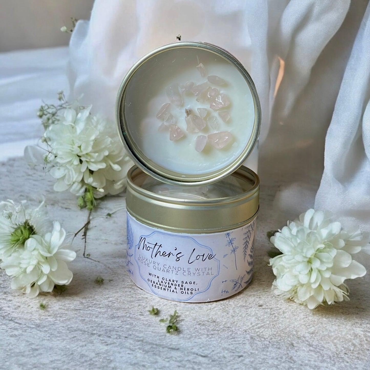 Candle in a tin with 'Mother's Love' label surrounded by white flowers on a light background