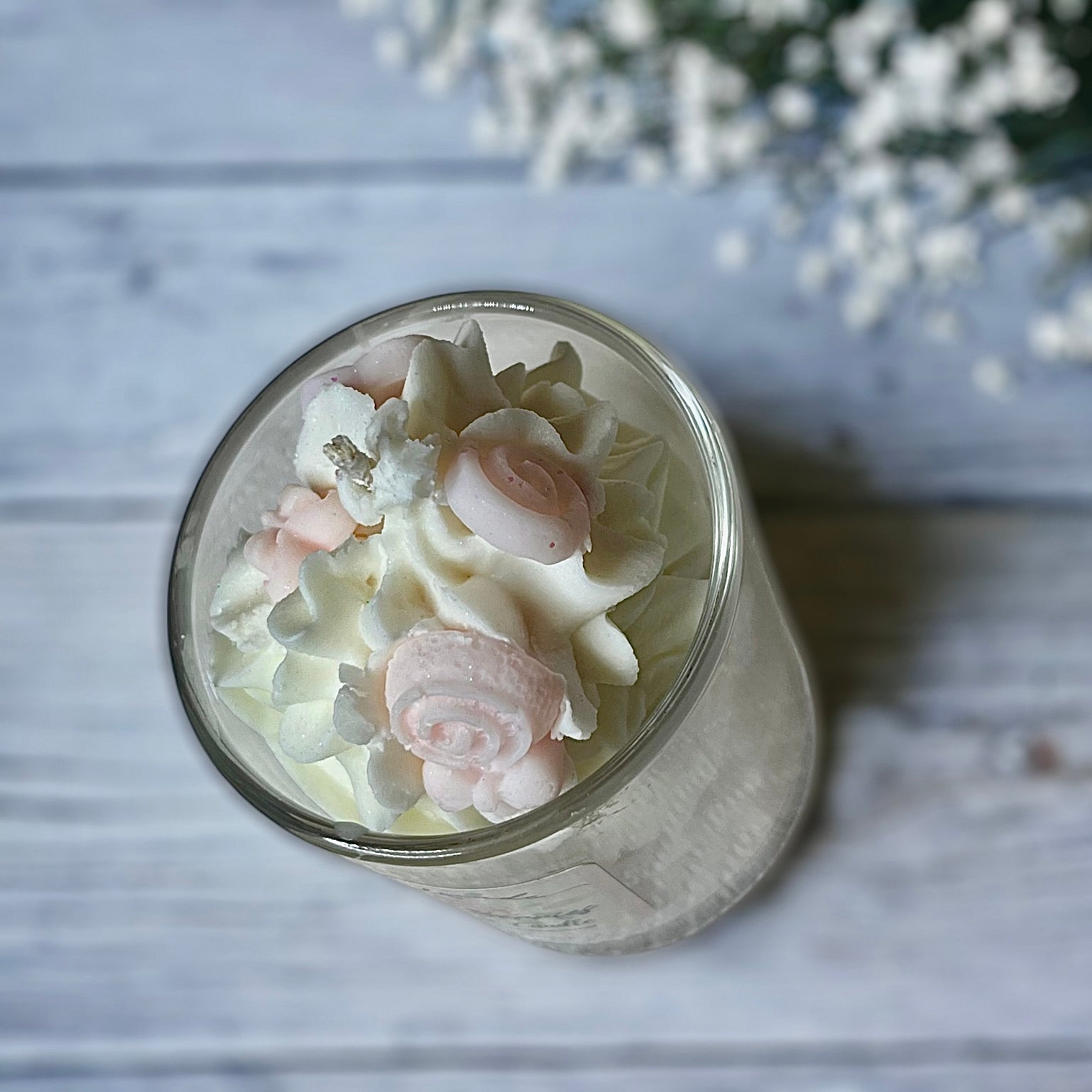 A clear glass candle with whipped wax topped with a white layer, decorated with pink and white heart-shaped wax melts, placed on a white surface with a background of white wood and flowers.
