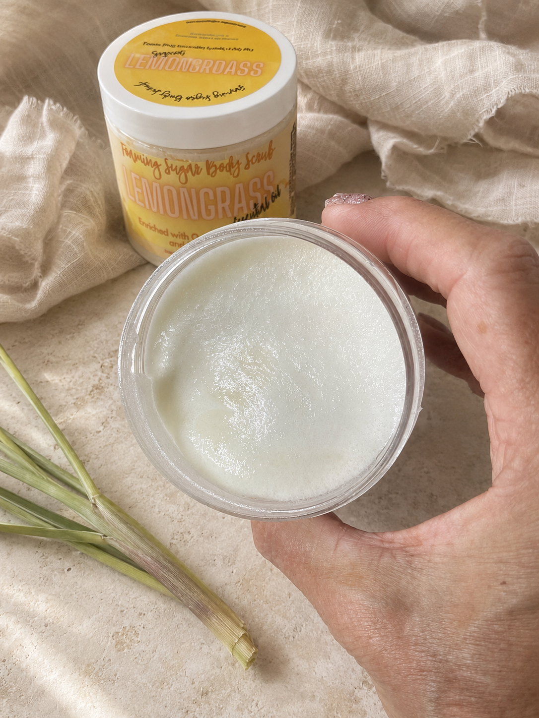 A hand holds an open clear plastic jar filled with a white creamy body scrub, with the branded round tub standing behind it on a beige fabric surface with linen and green stems as props.