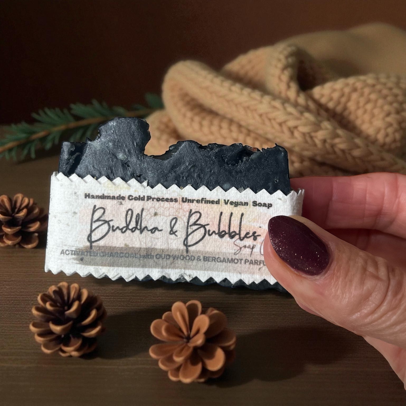 Handmade soap bar with 'Buddha & Bubbles' branding held by a hand, surrounded by pine cones and a textured fabric background.