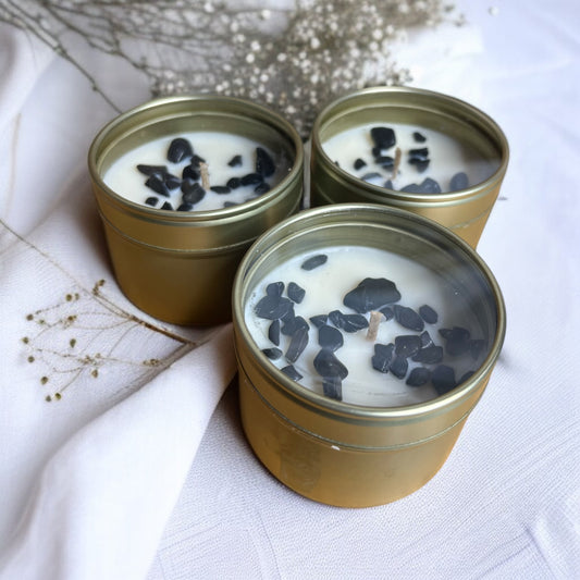 Three gold tins containing candles with black tourmaline crystals  on a white fabric background