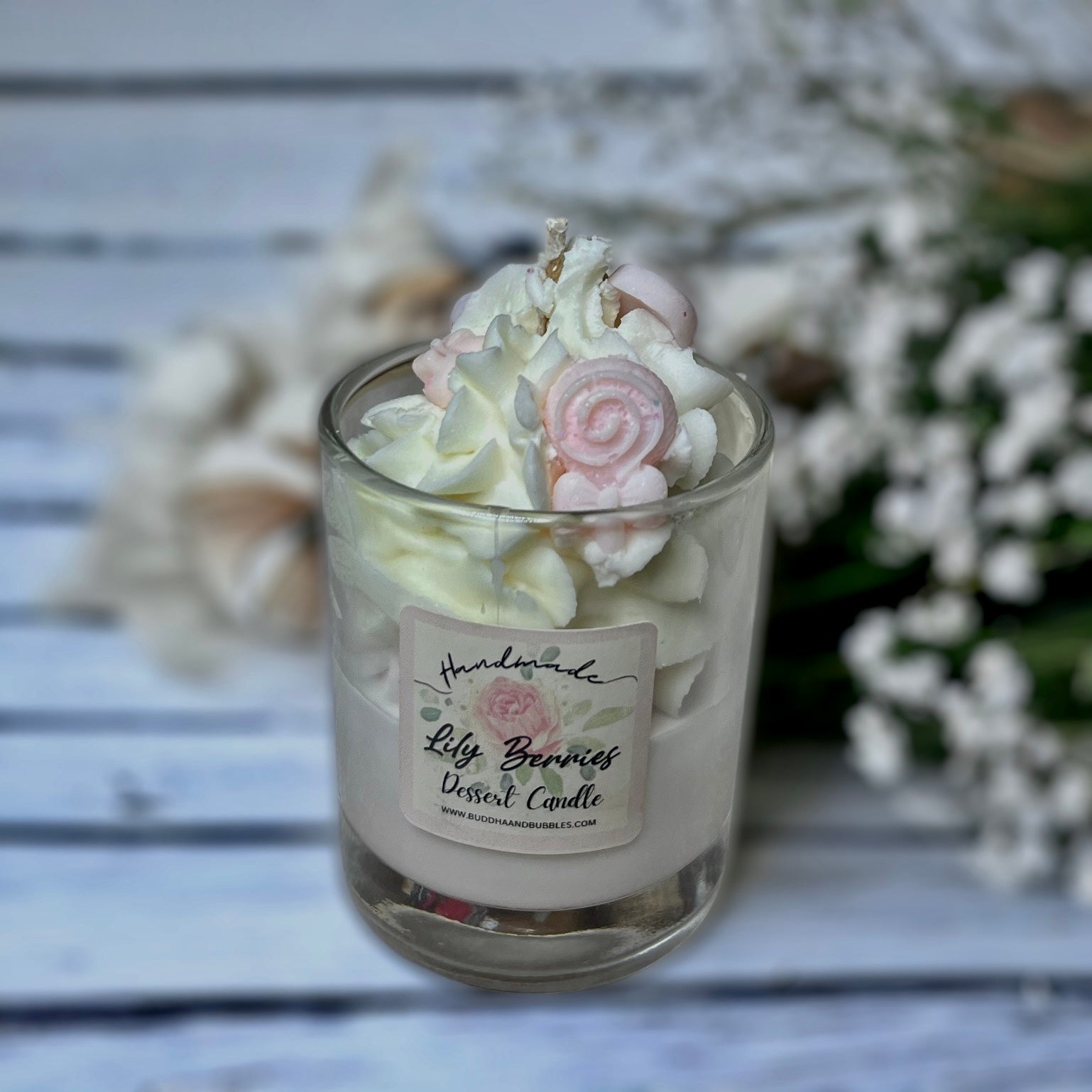 A clear glass candle with whipped wax topped with a white layer, decorated with pink and white heart-shaped wax melts, placed on a white surface with a background of white wood and flowers.
