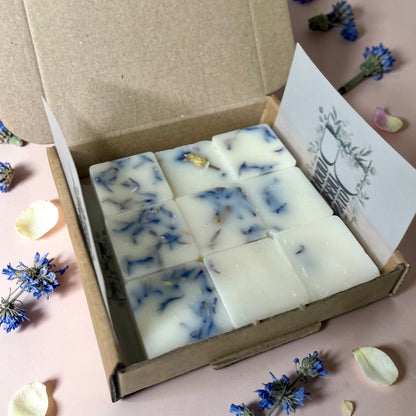 Box of white essential oil wax melts with blue mallow on a pink surface with flowers around.