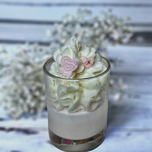 A clear glass candle with whipped wax topped with a white layer, decorated with pink and white heart-shaped wax melts, placed on a white surface with a background of white wood and flowers.