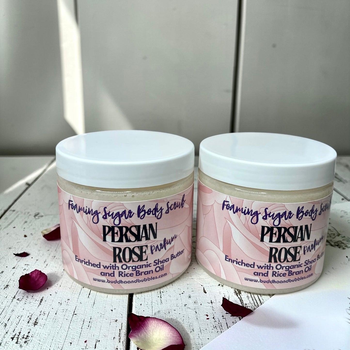 Two jars of Foaming Sugar Body Scrub in the Persian Rose Parfum scent, displayed side-by-side on a white wooden surface with scattered rose petals.