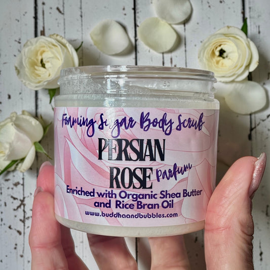 A hand holding a clear plastic jar of Buddha and Bubbles Foamng Sugar Body Scrub in Persian Rose Parfum. The jar contains a thick, semi-translucent off-white scrub, and the label is pink and white with purple text.