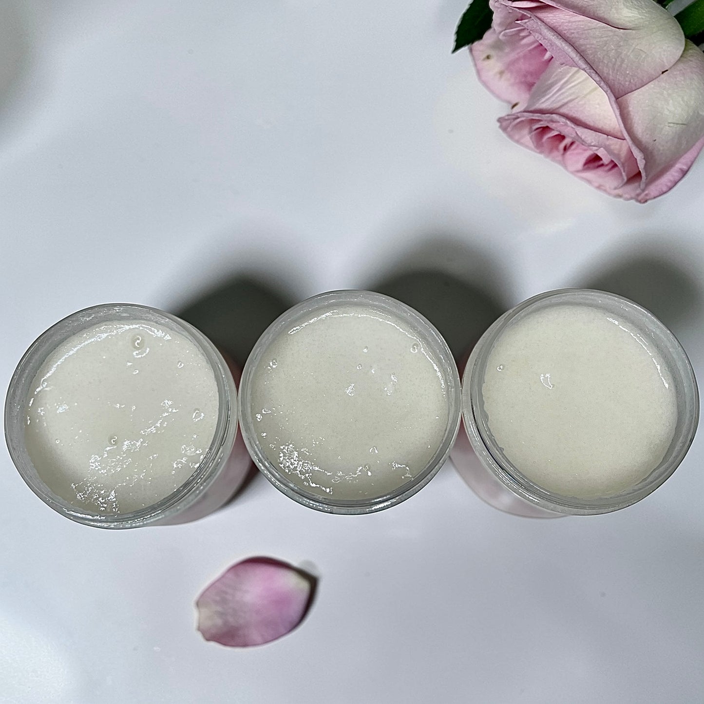 Three clear jars filled with a thick white sugar scrub arranged on a white surface with a pink rose petal and part of a pink rose in the background.