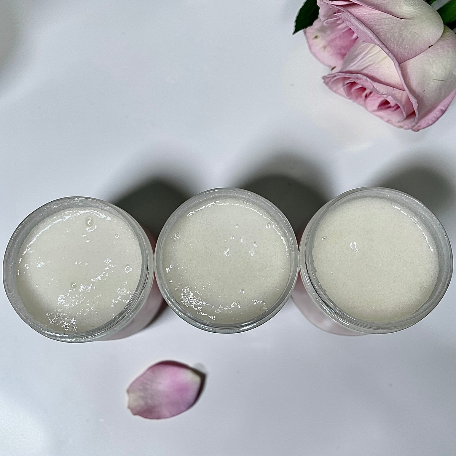 Three clear jars filled with a thick white sugar scrub arranged on a white surface with a pink rose petal and part of a pink rose in the background.