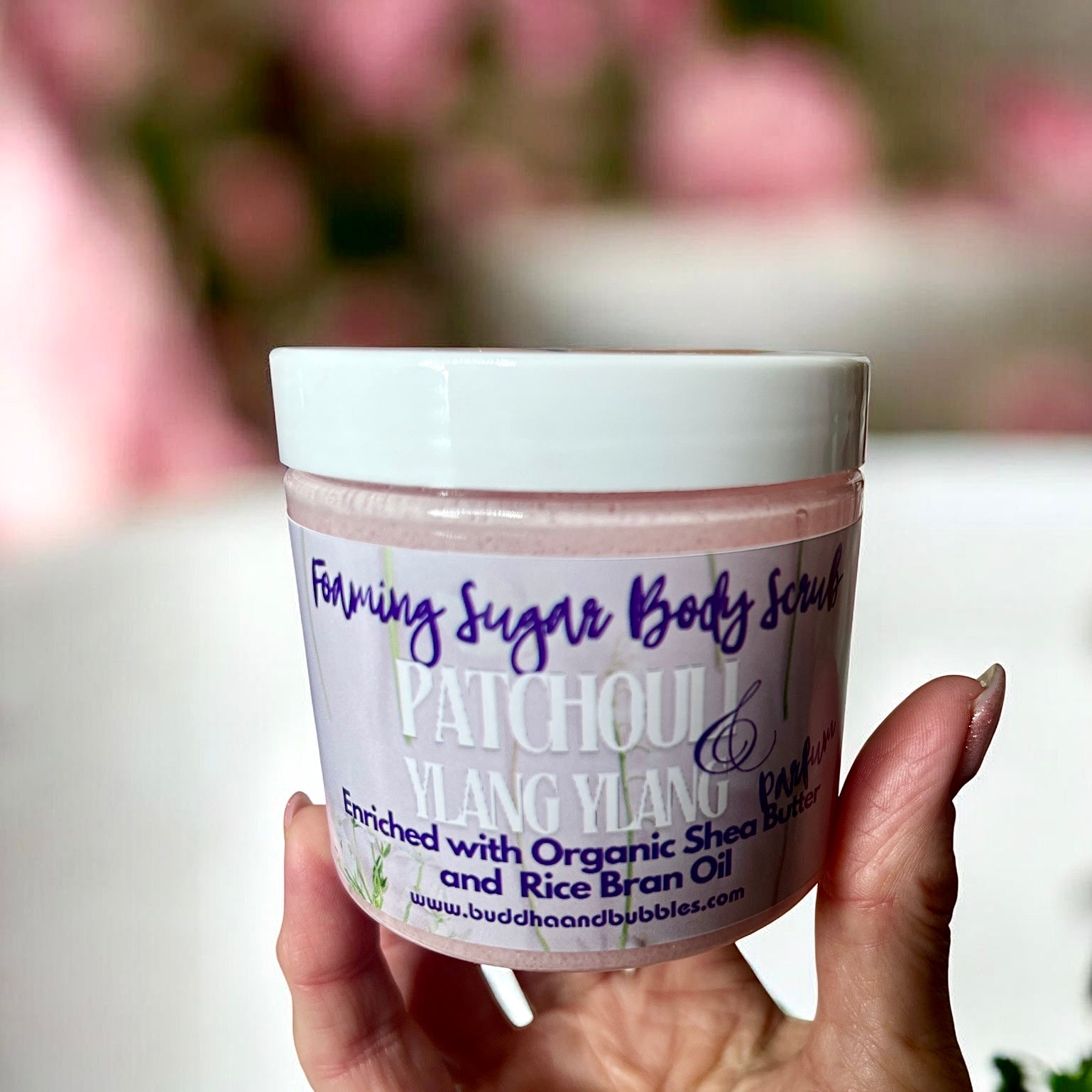 A hand holding a jar of foaming Sugar Body Scrub, Patchouli & Vanilia, with a white lid and a lavender label, set against a soft, blurred pink background.