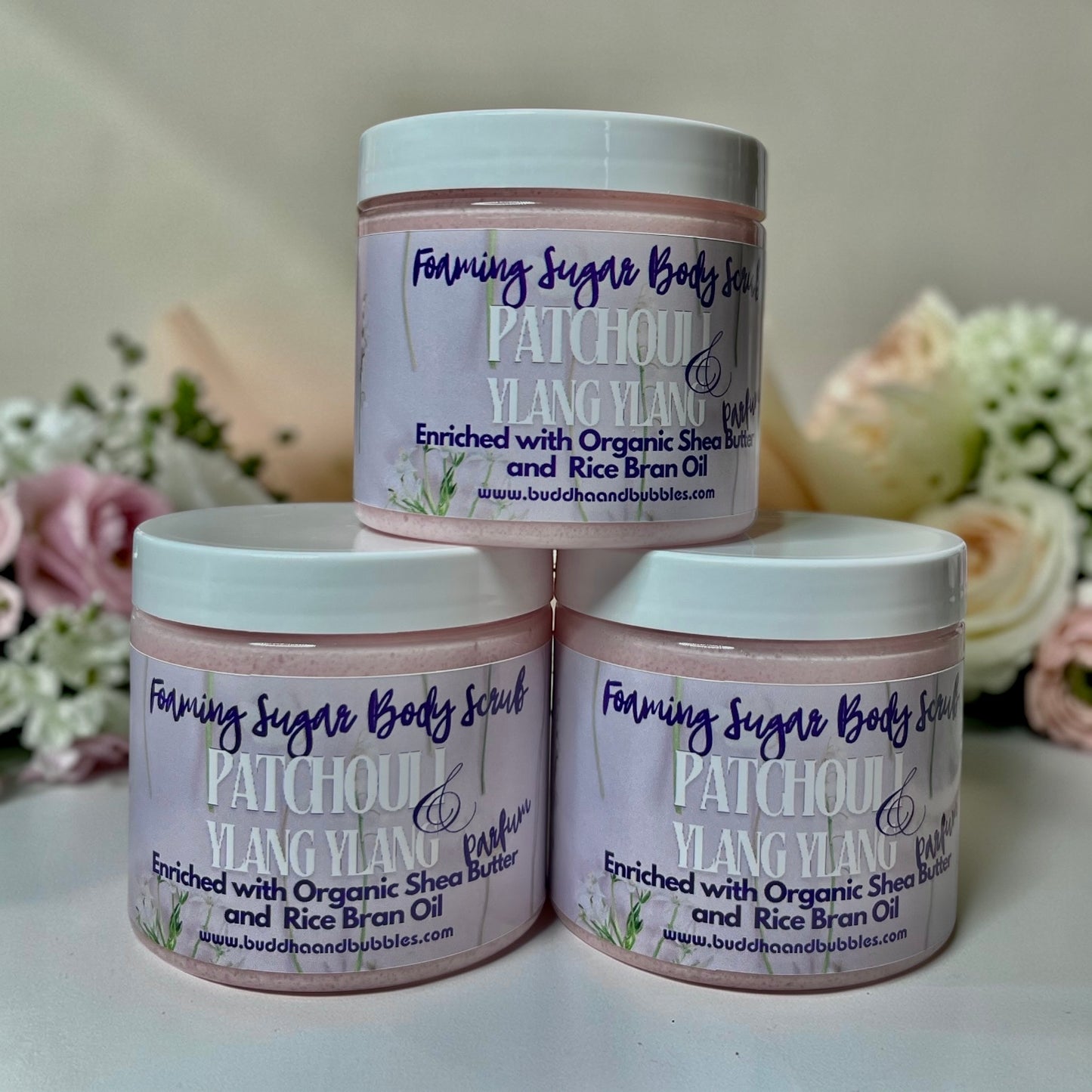 Three jars of Foaming Sugar Body Scrub Patchoul and Ylang Ylang enriched with Organic Shea Butter and Rice Bran Oil, arranged in a pyramid on a white surface with soft pink and white flowers in the background.