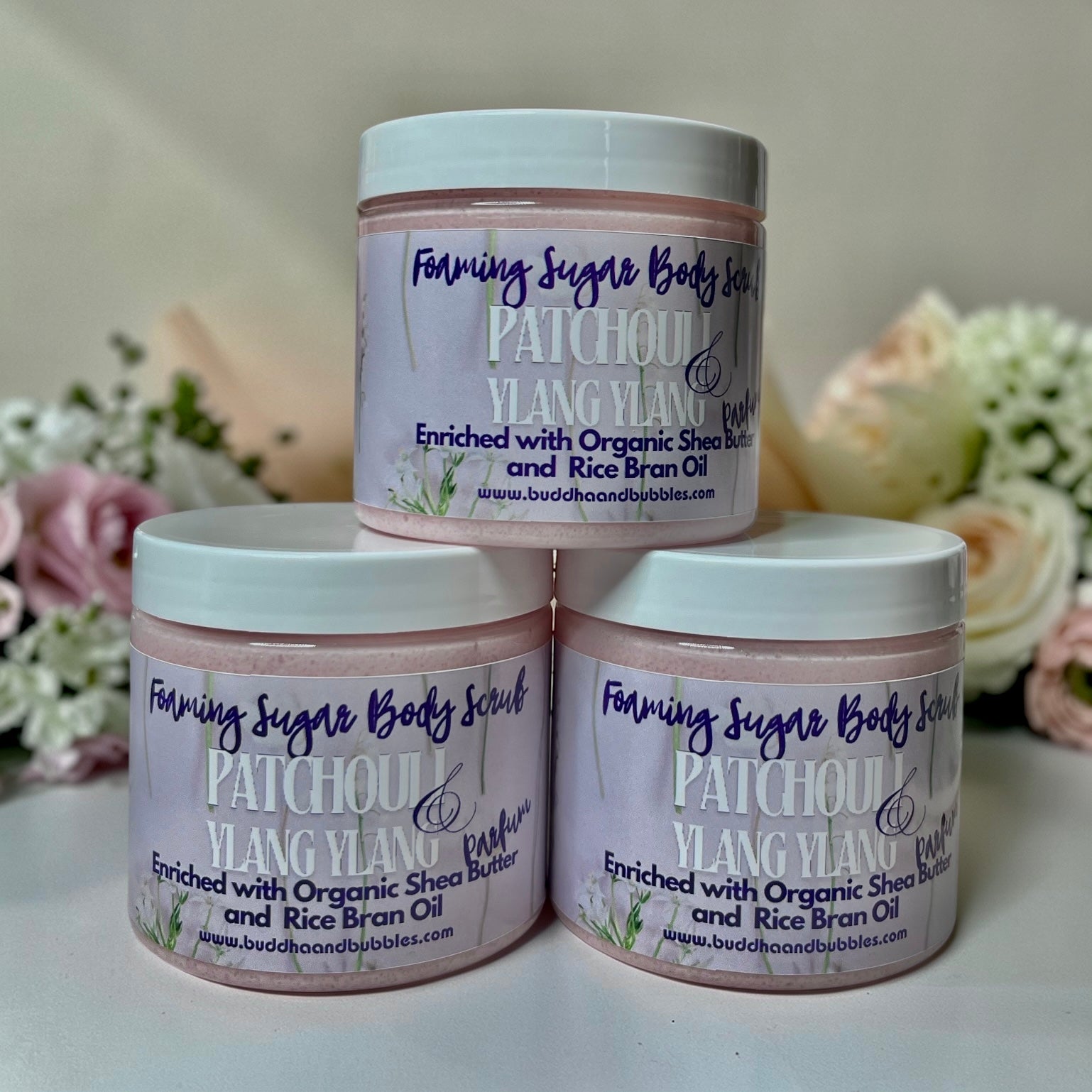 Three jars of Foaming Sugar Body Scrub Patchoul and Ylang Ylang enriched with Organic Shea Butter and Rice Bran Oil, arranged in a pyramid on a white surface with soft pink and white flowers in the background.