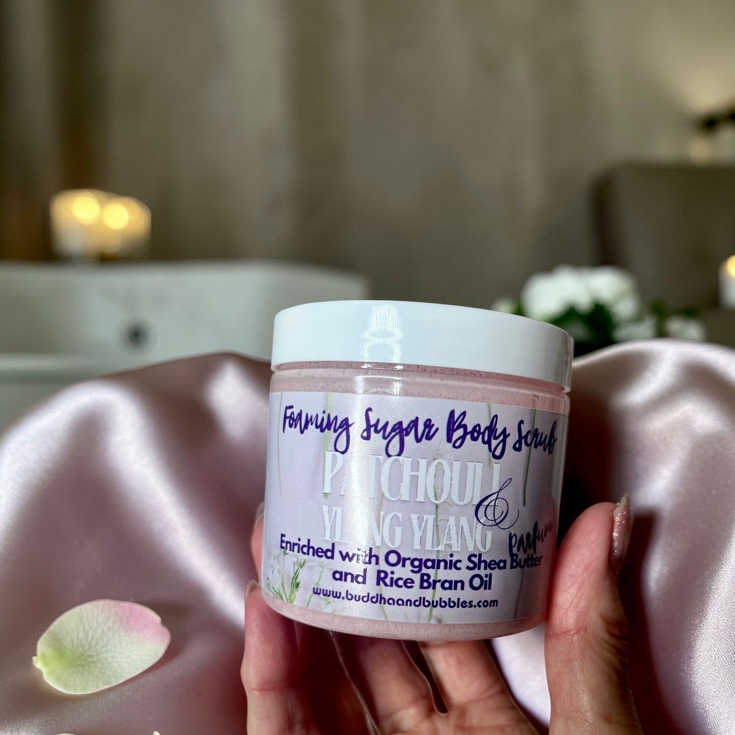 A hand holding a jar of foaming Sugar Body Scrub by Patchouli & Ylang Ylang featuring a white lid and a lavender label with purple text, set against a soft pink satin fabric background.