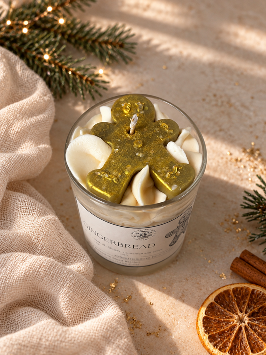 A glass jar candle labeled Gingerbread, filled with white wax and topped with a gold gingerbread figure, styled on a beige surface with pine branches, cinnamon sticks, and a dried orange slice.