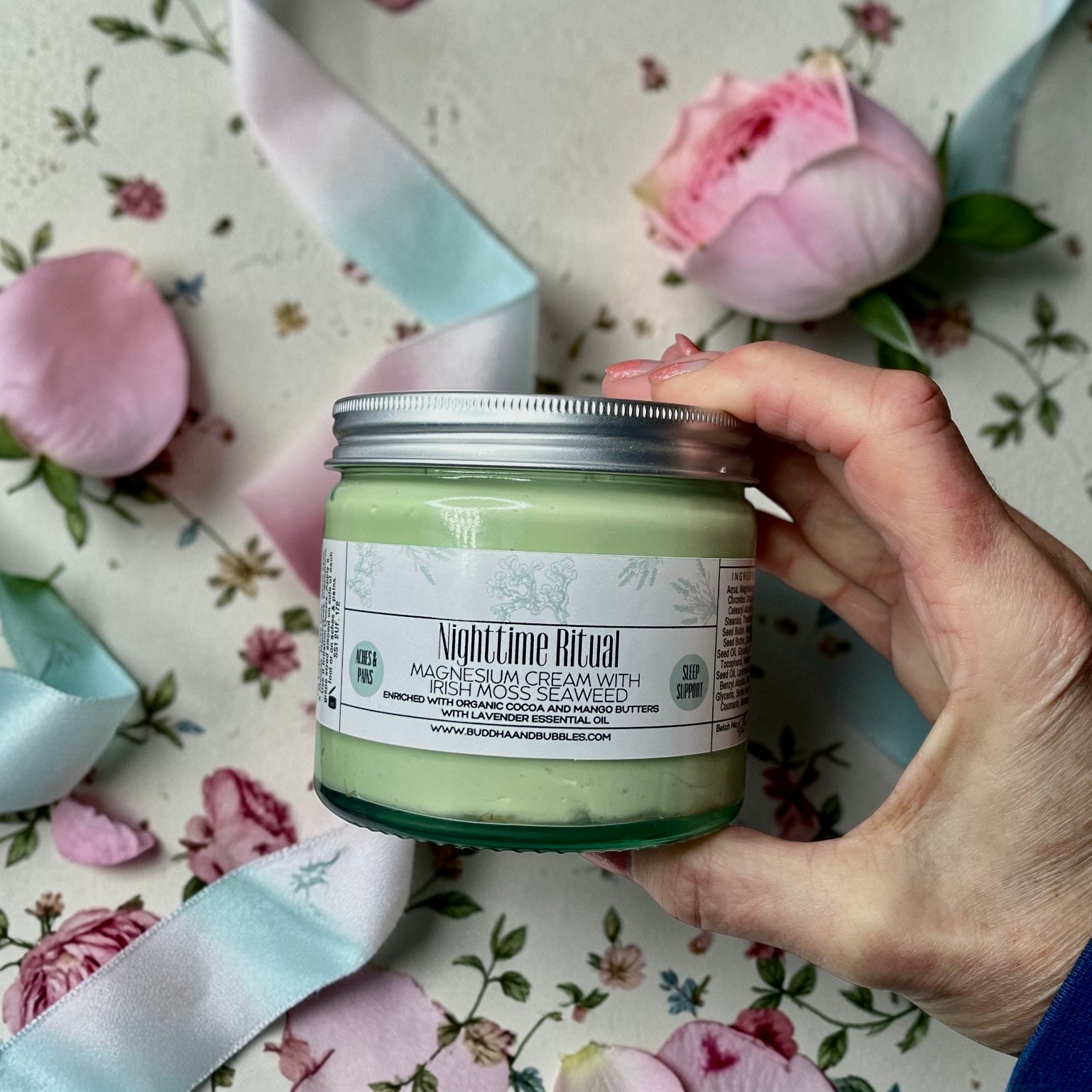 Hand holding a jar of jumbo size jar of 'Nighttime Ritual' magnesium  cream with flowers and ribbons in the background