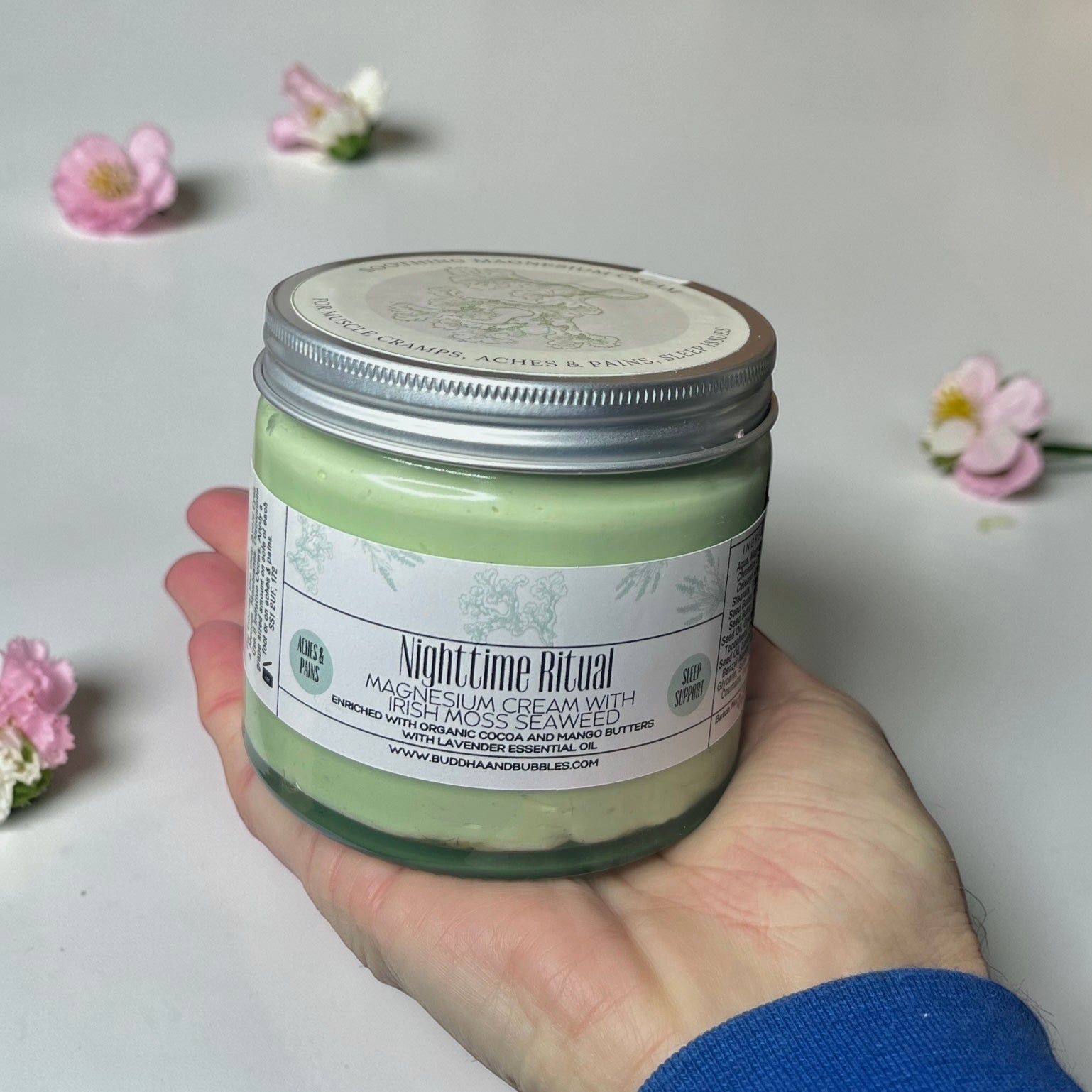 Hand holding a jumbo jar of 'Nighttime Ritual' magnesium cream with flowers in the background