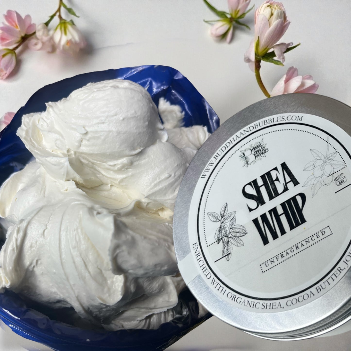 Shea whip body butter in a tin container with a silver lid on a white background with flowers.