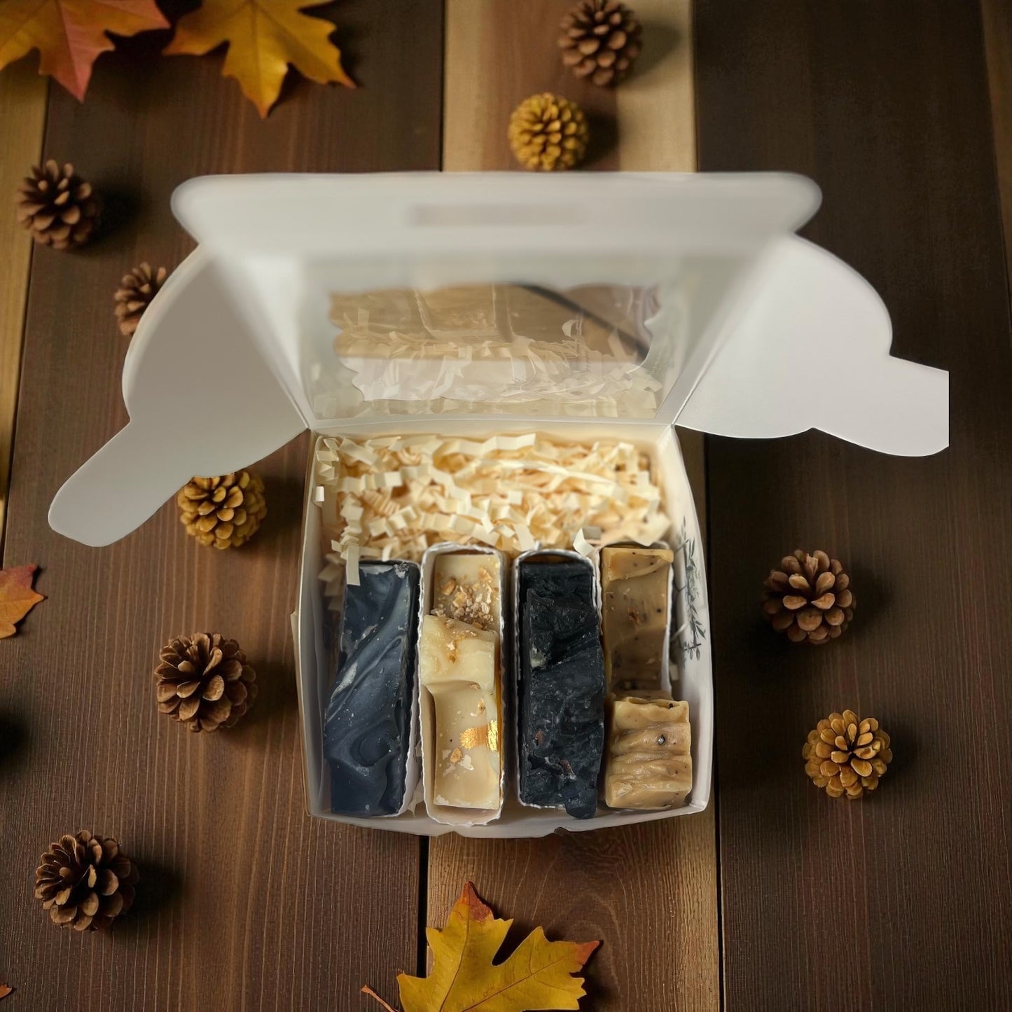 Assorted soaps in a white gift box on a wooden surface with pinecones and leaves.