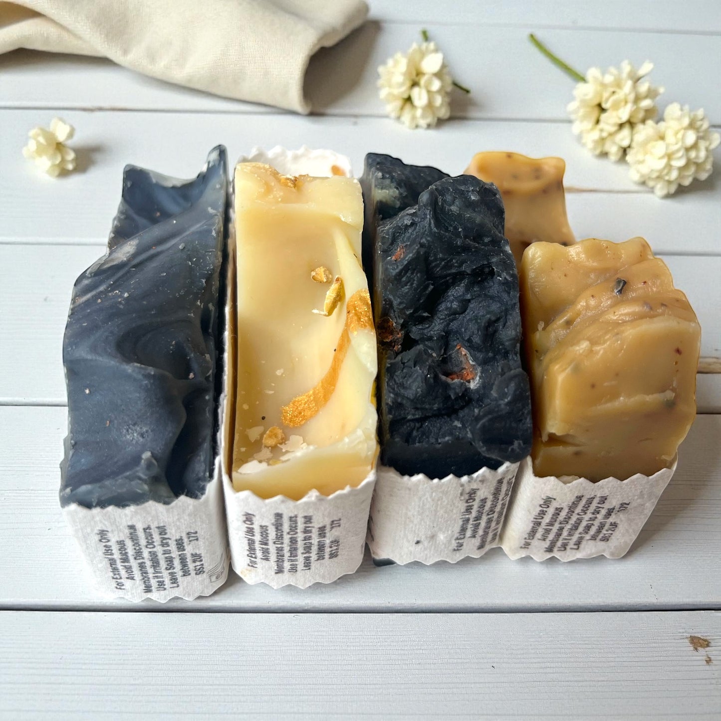 Four bars of soap in various colours and textures on a white wooden surface with flowers.