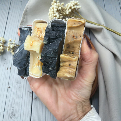 Hand holding three different types of soap bars on a wooden surface with dried flowers.