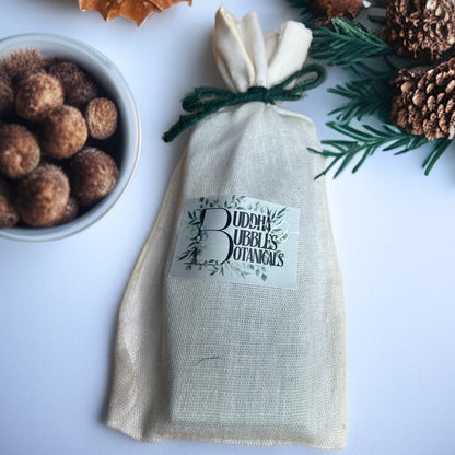 cotton reusable bag containing a wax melt snap bar with 'Buddha Bubbles Botanicals' label next to a bowl of brown nuts and pinecones.