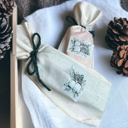 Three small fabric bags containing wax melt snap bar with labels on a white surface with pine cones.