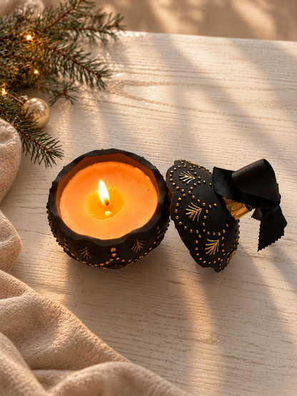 A black decorative christmas bauble candle with gold patterns and a matching lid tied with a black ribbon, shown beside a lit orange candle on a light wood surface with holiday greenery and a beige blanket nearby.