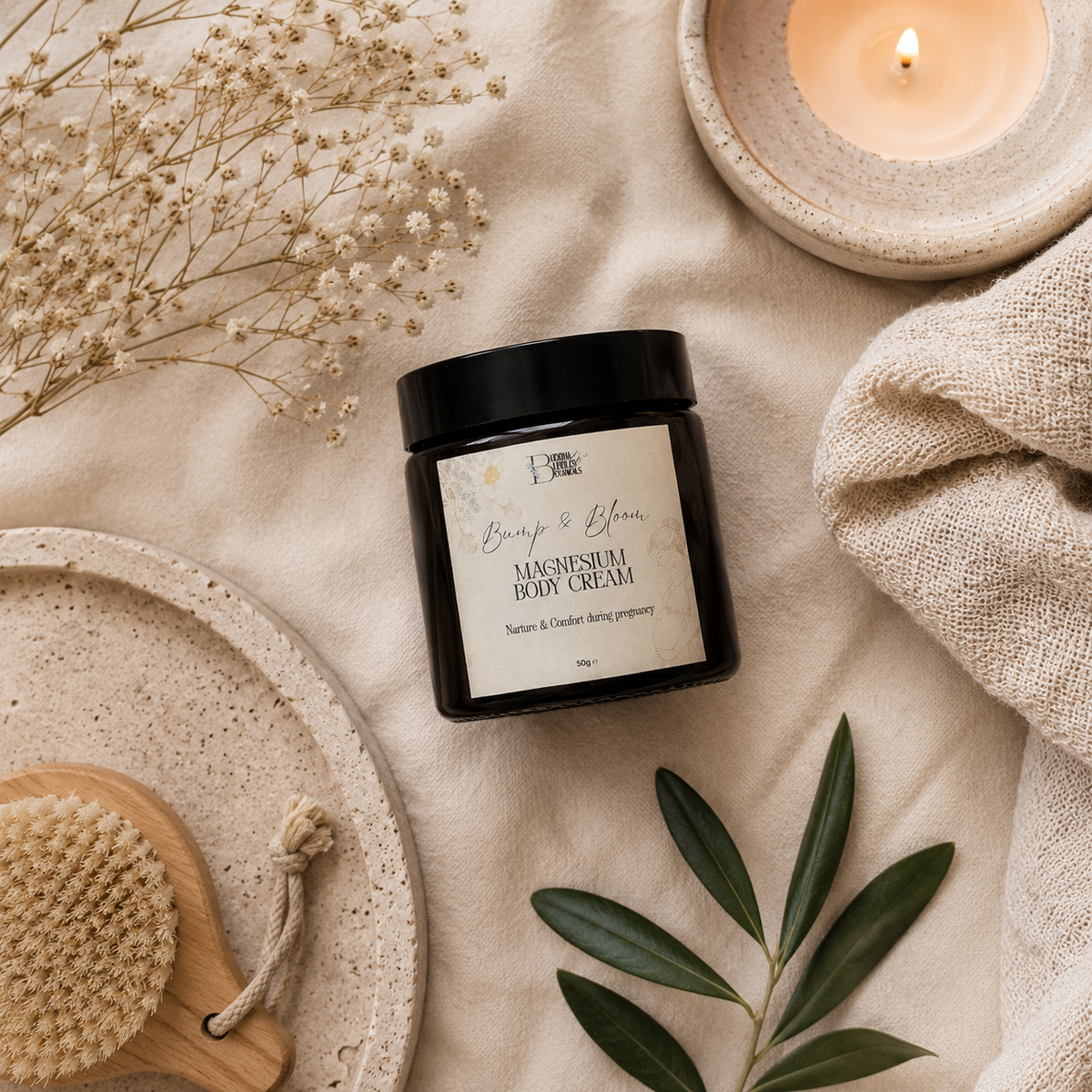 A jar of Beauty & Bloom Magnesium Body Cream is styled in a flat lay on a beige linen fabric with a lit candle, dried flowers, a wooden brush, and green leaves.