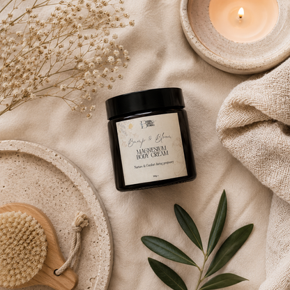 A jar of Beauty & Bloom Magnesium Body Cream is styled in a flat lay on a beige linen fabric with a lit candle, dried flowers, a wooden brush, and green leaves.