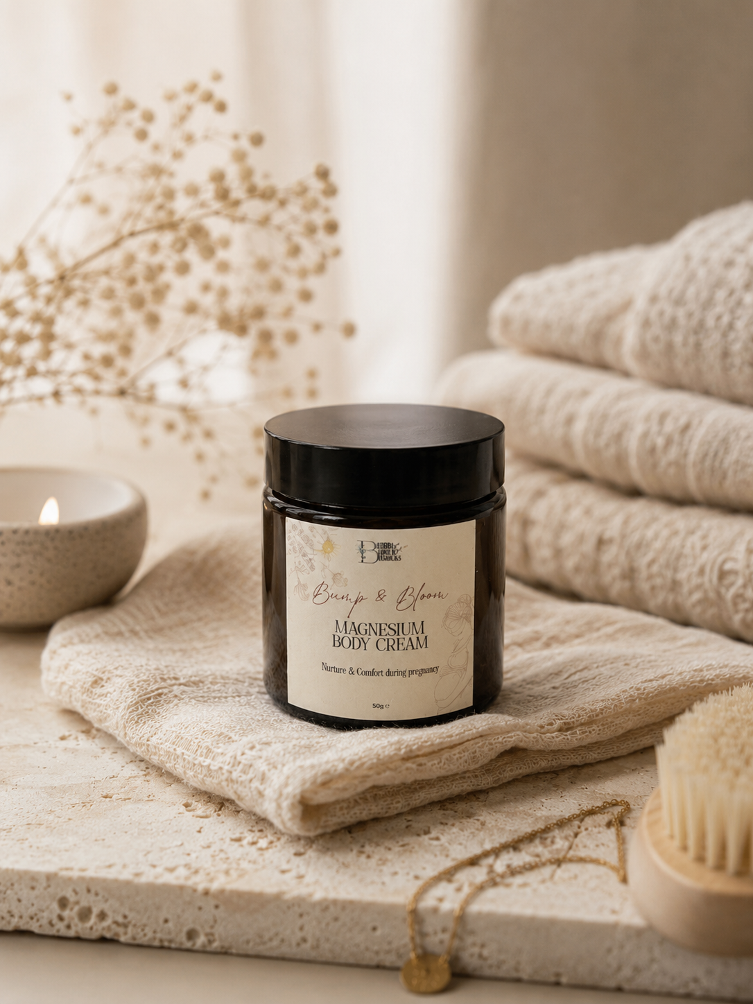 A jar of Bump & Bloom Magnesium Body Cream sits on a textured cream towel, surrounded by spa props including folded towels, a candle, dried flowers, and a wooden brush.
