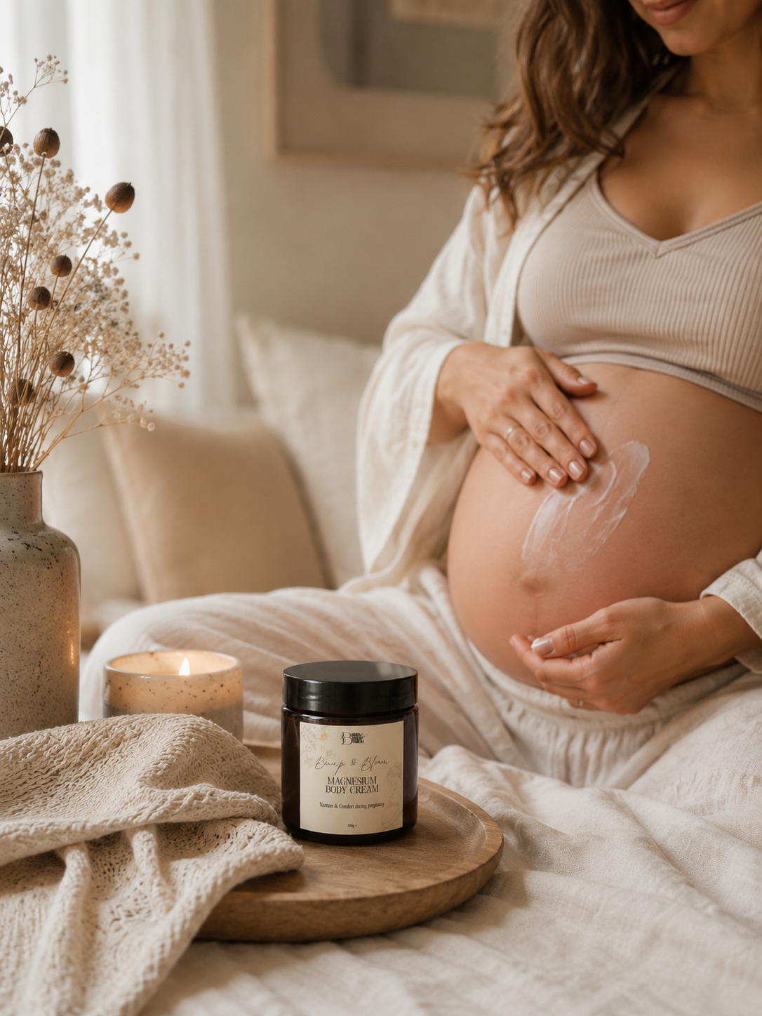 A pregnant woman in a cozy bedroom setting applies Bump & Bloom Magnesium Cream to her belly while a jar of Bump and Bloom Magnesium Cream sits on a wooden tray in the foreground