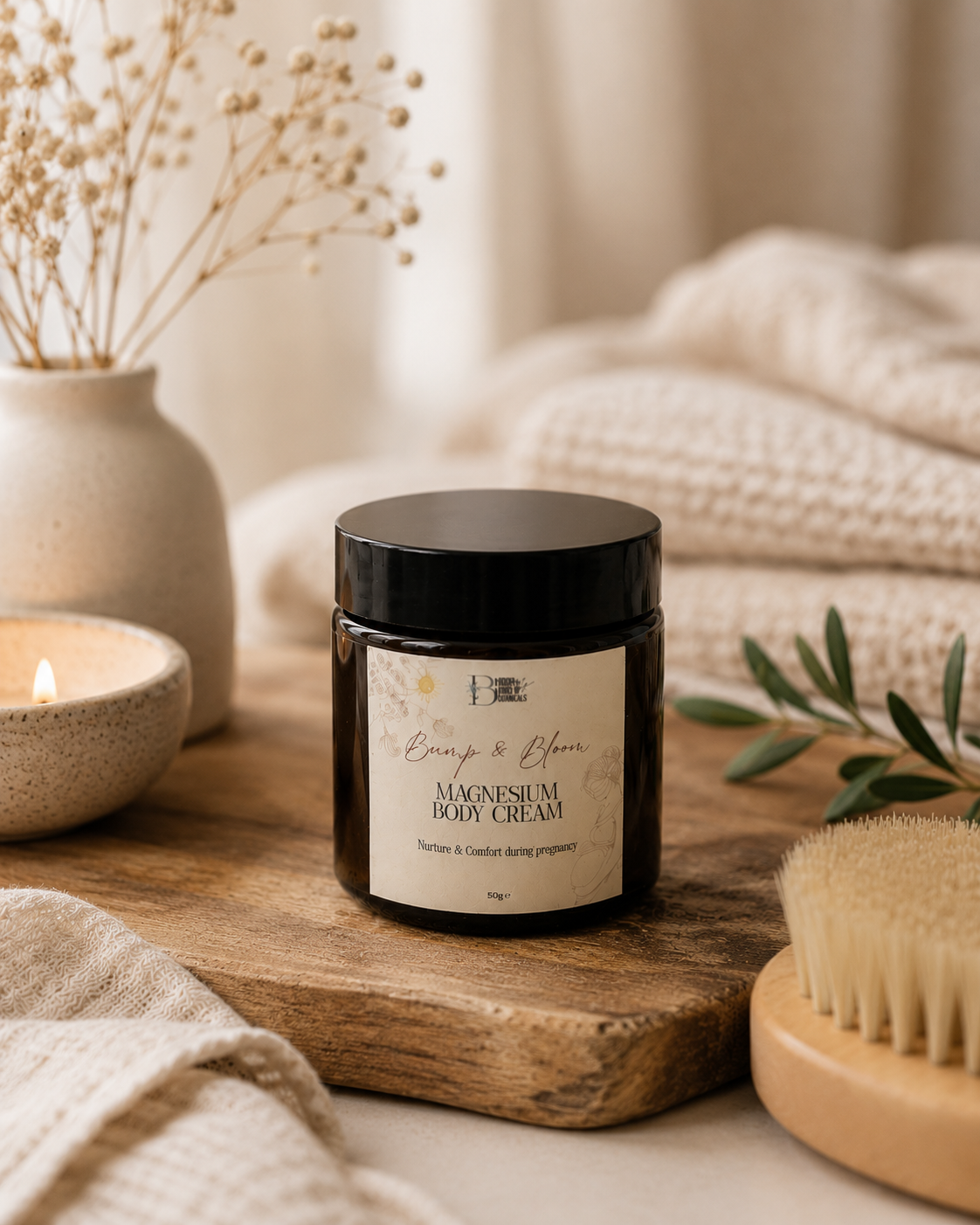 A jar of Bump & Bloom Magnesium Body Cream sits on a rustic wooden board surrounded by cozy bath-themed props, including a lit candle, a woven towel, dried flowers, and a natural brush.