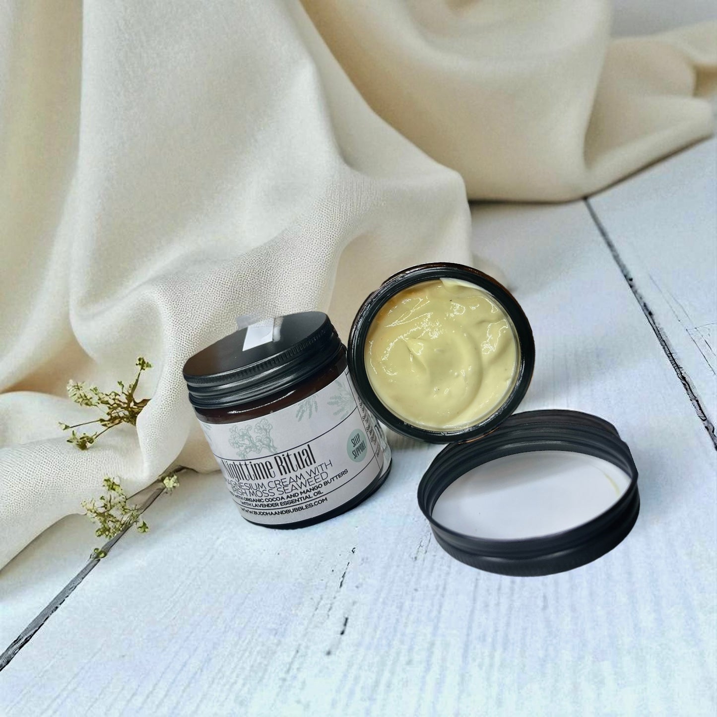 Two jars of cream, one open showing a white magnesium cream, on a light wooden surface with a beige muslin cloth in the background.