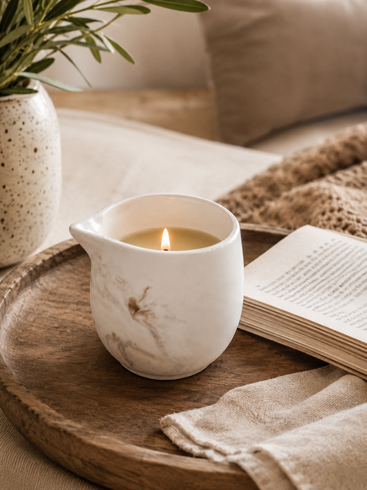 massage Candle in a white ceramic holder on a wooden tray with a book and plant in the background